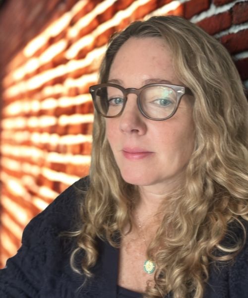 white woman with curly hair navy seater and glasses in front of brick wall