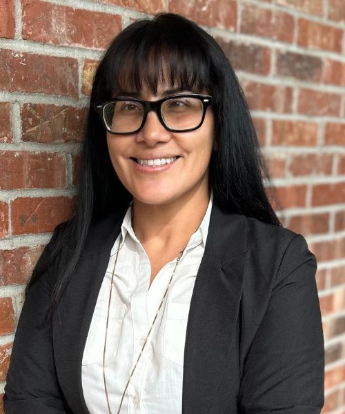 woman, black hair, blazer, smiling in front of brick wall